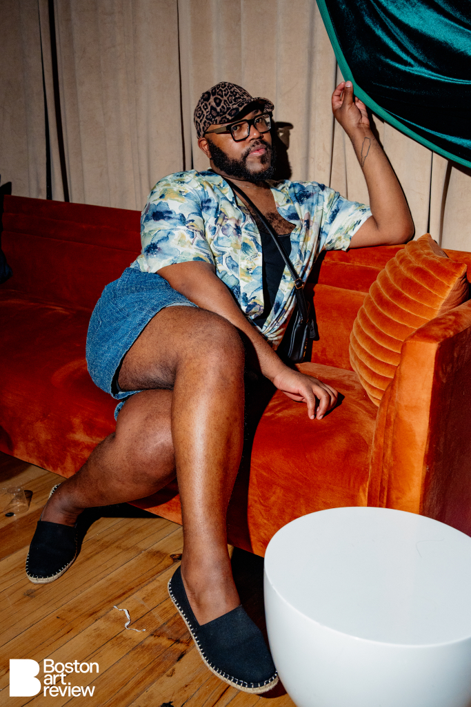 Sheldon Gray of Dive Bar Caviar seated on Kadeema Rentals' Atlas Sofa at the Boston Art Review Issue 14 launch party, featuring the White Tam Tam Table, velvet accent pillow, and luxury drapery by Simplistic Charm.