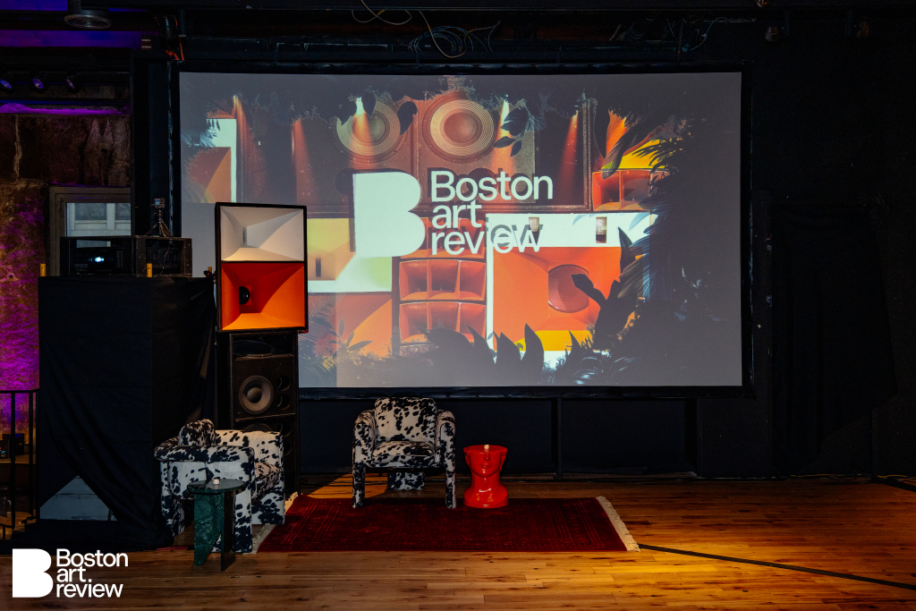 Kadeema Rentals lounge setup at the Boston Art Review Issue 14 launch party featuring black and white cowhide-print chairs, a red sculptural side table, a green marble accent table, and a layered rug in front of a Boston Art Review projection screen.