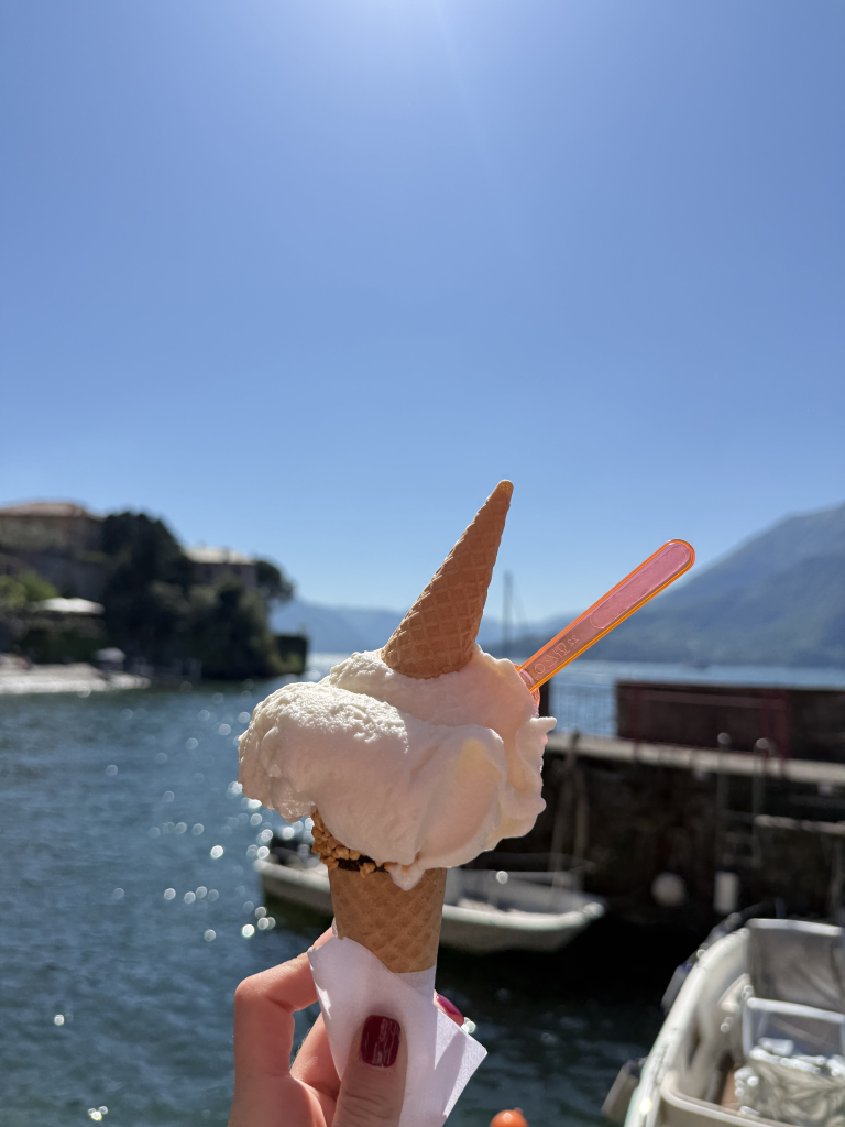 Gelato cone held up beside Lake Como with the water and mountains in the background, Italy